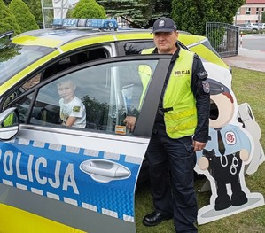 Policjant na tle oznakowanego radiowozu, wewnątrz, którego  siedzi dziecko