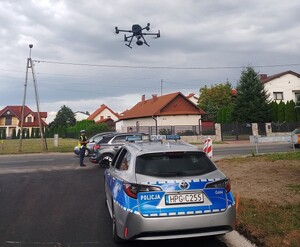 Startujący dron policyjny w otoczeniu oznakowanego radiowozu. Na drugim planie stoi policjant ruchu drogowego