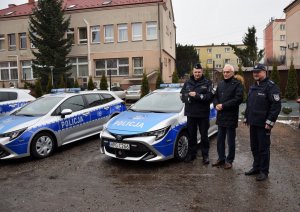 Burmistrz Szczucina wraz z Komendantem powiatowym Policji oraz Komendantem Komisariatu Policji w Szczucinie stoją obok radiowozu, Komendant Komisariatu w rękach trzyma dokumenty i kluczyki do samochodu służbowego