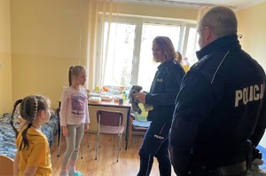 policjantka trzyma misia policjant stoi obok, dwie dziewczynki w pomieszczeniu internatu w tle stół i łóżko