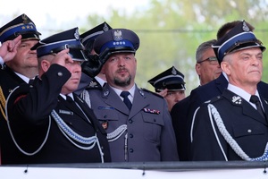 Komendant Powiatowy Policji ubrany na galowo oddaje honor, obok z dwóch stron strażacy osp ubrani również na galowo