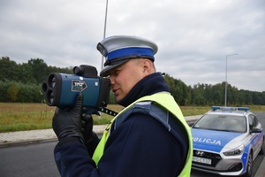 Zdjęcie przedstawia umundurowanego policjanta mierzącego prędkość za pomocą urządzenia pomiarowego